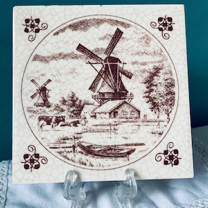 Può includere: Piastrella quadrata in ceramica con un'illustrazione color seppia di una scena di mulini a vento olandesi. L'immagine presenta mulini a vento, una casa, una barca e una mucca. Accenti floreali decorativi agli angoli incorniciano l'immagine centrale. La piastrella è esposta su un supporto di plastica trasparente.