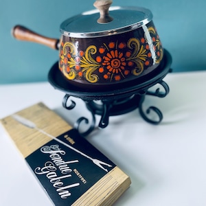 Può includere: Un pentolino per fondue vintage con una base marrone e un motivo floreale in arancione, giallo e verde. Il pentolino ha un coperchio argentato e un manico in legno. Il pentolino è appoggiato su un supporto in metallo nero. Una scatola di forchette per fondue è in primo piano. La scatola è marrone con testo bianco che dice "Fondue Gabeln Rostfrei!"