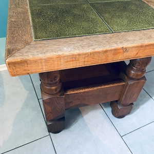 Puede incluir: Una mesa de café cuadrada de madera con una tapa de azulejos verdes y cuatro patas torneadas. La mesa tiene un estante inferior.