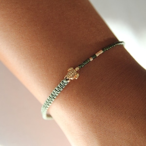 Könnte beinhalten: Nahaufnahme eines Armbands mit einem goldfarbenen Schildkrötenanhänger. Das Armband hat ein geflochtenes, hellgrünes Band mit dunkelgrünen und goldfarbenen Perlen. Das Armband ist für das Handgelenk konzipiert.