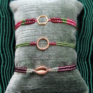 Könnte beinhalten: Drei zarte Armbänder mit goldenen Akzenten auf einem grauen Samtkissen. Die Armbänder bestehen aus kleinen grünen und rosa Perlen mit einem Hexagon, einem Kreis und einem Muschel-Anhänger.
