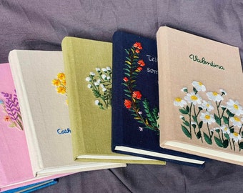 Può includere: Cinque diari rilegati con diversi disegni floreali. I diari sono beige, verde, blu e rosa. I nomi "Cath", "Tel", "Some" e "Valentina" sono ricamati sulle copertine.