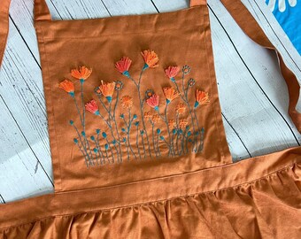 Puede incluir: Un delantal de lino marrón con un diseño de bordado floral. El bordado presenta flores naranjas y azul verdoso con tallos verdes.