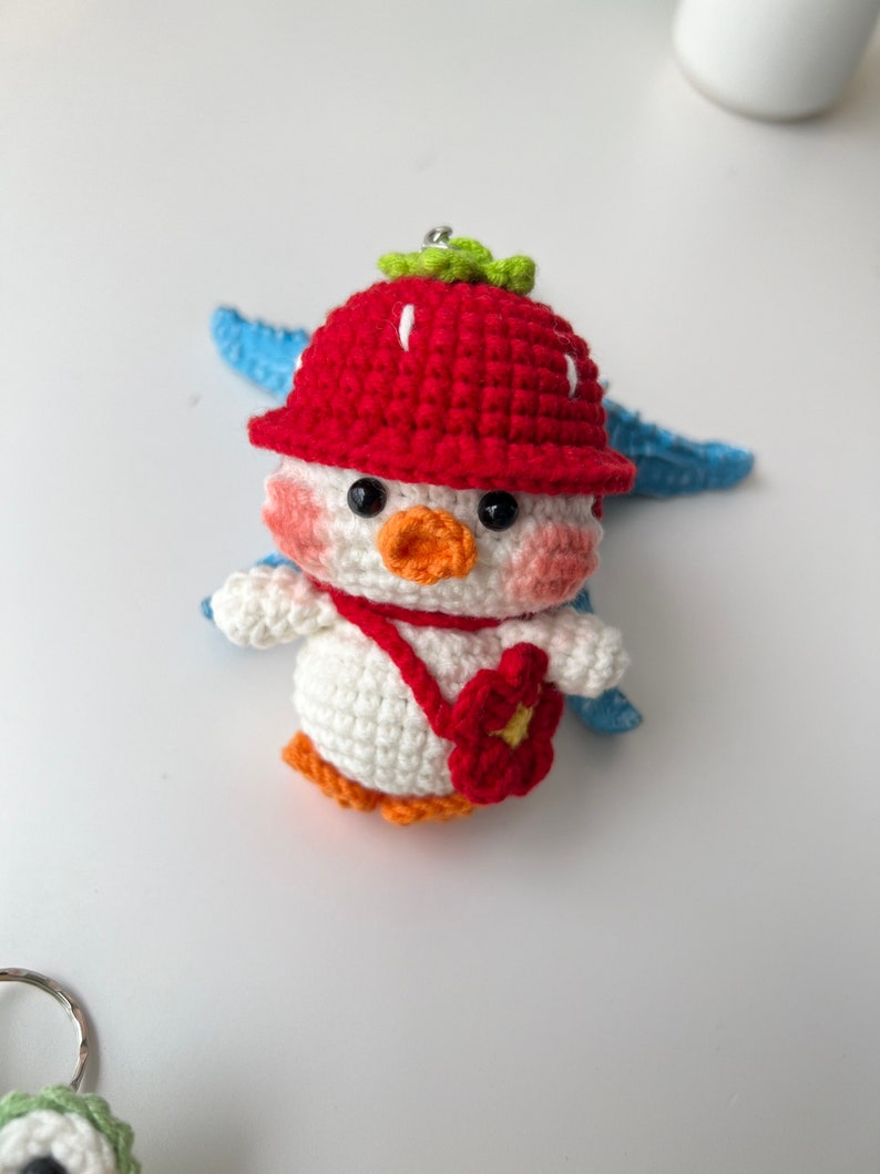 May include: A white crocheted penguin wearing a red strawberry hat with green leaves and a red flower purse. The penguin has black eyes, an orange beak, and pink cheeks.