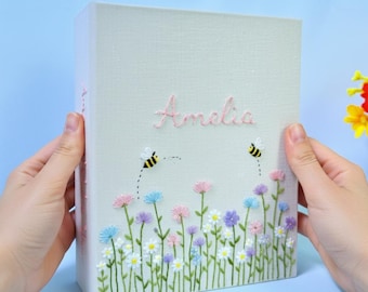 May include: A light-colored, rectangular box with embroidered floral and bee designs. The name "Amelia" is embroidered in pink cursive. The flowers are in various colors, including pink, blue, purple, and white.