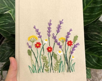 May include: A beige linen notebook with an embroidered design of purple lavender flowers, yellow and red wildflowers, and green leaves.