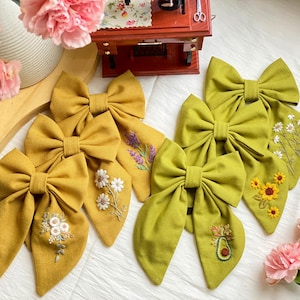 K&ouml;nnte beinhalten: F&uuml;nf handgefertigte Stoffhaarb&auml;nder mit gestickten Blumenmustern. Die Schleifen sind in zwei Farben gehalten: Senfgelb und Olivgr&uuml;n. Die Designs beinhalten Lavendel, G&auml;nsebl&uuml;mchen, Sonnenblumen und eine Avocado.