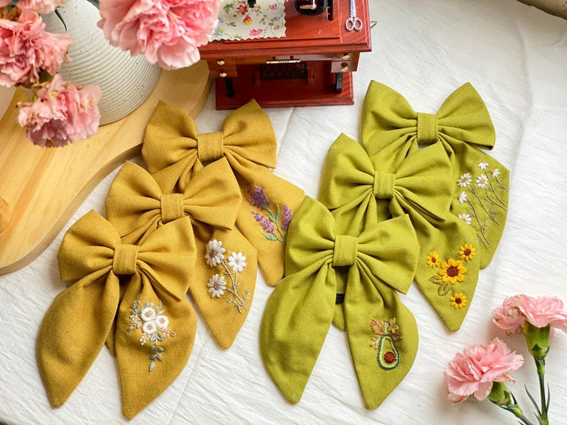 K&ouml;nnte beinhalten: F&uuml;nf handgefertigte Stoffhaarb&auml;nder mit gestickten Blumenmustern. Die Schleifen sind in zwei Farben gehalten: Senfgelb und Olivgr&uuml;n. Die Designs beinhalten Lavendel, G&auml;nsebl&uuml;mchen, Sonnenblumen und eine Avocado.