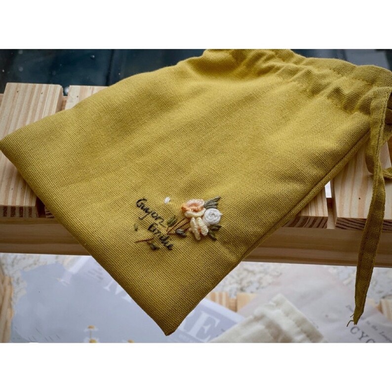 May include: A mustard yellow drawstring bag with a small embroidered floral design and the text "Gayer than Frida" in black thread.