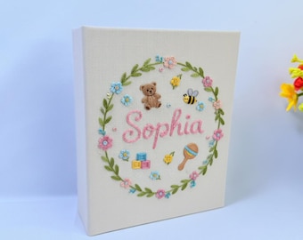 May include: A cream-colored baby keepsake box with an embroidered floral wreath design. The wreath includes a teddy bear, bee, rattle, and the name "Sophia" in pink. The box is a perfect gift for a baby.