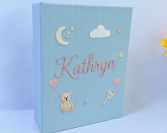 May include: A light blue baby keepsake box embroidered with the name "Kathryn" in pink script. The box features embroidered images of a moon, stars, clouds, hearts, a teddy bear, and a rattle. A charming and personalized baby gift.