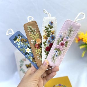 K&ouml;nnte beinhalten: Vier bestickte Stoff-Lesezeichen in Blau, Braun, Wei&szlig; und Rosa. Jedes Lesezeichen hat ein anderes Blumendesign mit einer Schlaufe oben. Die Lesezeichen werden in einer Hand gehalten.