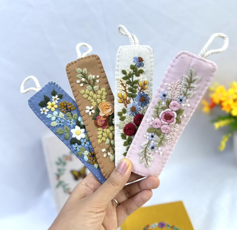 K&ouml;nnte beinhalten: Vier bestickte Stoff-Lesezeichen in Blau, Braun, Wei&szlig; und Rosa. Jedes Lesezeichen hat ein anderes Blumendesign mit einer Schlaufe oben. Die Lesezeichen werden in einer Hand gehalten.