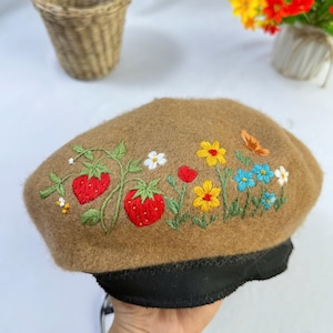 May include: A tan beret with colorful floral embroidery, including red strawberries, yellow, blue, and white flowers, and a butterfly. The beret has a black band at the base.