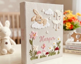 May include: A beige fabric-covered photo album featuring intricate embroidery of a sleeping bunny on a cloud, a crescent moon, flowers, mushrooms, and a butterfly. The name Harper is embroidered in pink, and the year 2026 is on the spine.