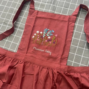 Puede incluir: Un delantal marrón con un diseño de bordado floral y el texto "Mommom Debby". El delantal tiene un dobladillo con volantes y un bolsillo grande.