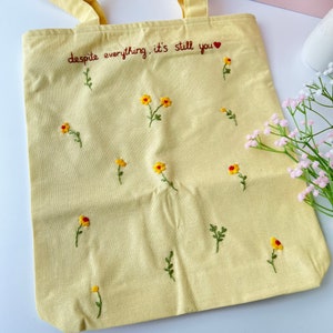May include: A yellow canvas tote bag with the embroidered text "despite everything, it's still you" and small yellow flowers.