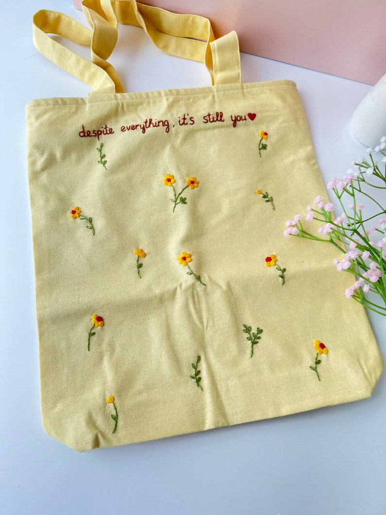 May include: A yellow canvas tote bag with the embroidered text "despite everything, it's still you" and small yellow flowers.