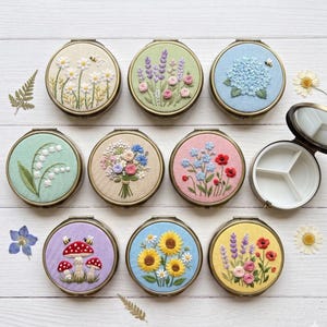Pode incluir: Uma coleção de oito porta-comprimidos redondos de metal com tampas de tecido bordado com desenhos florais, botânicos e de cogumelos. Um estojo aberto revela um interior de plástico branco com três compartimentos. Cada tampa tem motivos únicos.