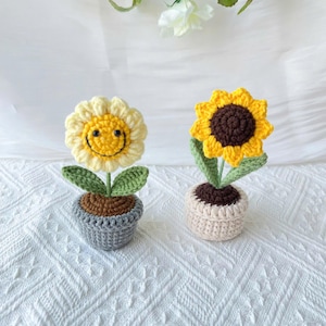 May include: Two crocheted sunflower plants in pots. One sunflower has a smiley face and the other has a brown center. Both are on a white background.