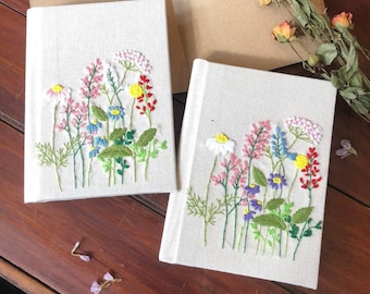Puede incluir: Dos diarios de lino blanquecino con diseños florales bordados. El colorido bordado presenta flores rosas, azules, amarillas y rojas con hojas verdes. Los diarios están abiertos sobre una superficie de madera.