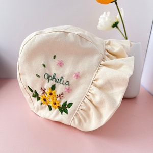 May include: A cream-coloured sun hat with a ruffled edge and embroidered flowers and the name "Ophelia" in green thread.