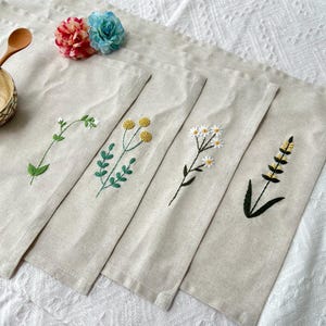 Könnte beinhalten: Vier beigefarbene Leinen-Tischsets mit gestickten Blumenmustern. Jedes Set zeigt eine andere Blume: einen Zweig mit weißen Blüten, gelbe Pompons, weiße Gänseblümchen und eine gelb-schwarze Ähre. Ein Holzlöffel und eine Schüssel sind sichtbar.