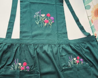 May include: A dark green apron with embroidered flowers in shades of pink, purple, and white. The apron has two pockets on the front.