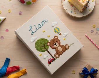 May include: A cream-colored, embroidered book cover with the name "Liam" in blue script. The cover features a brown teddy bear, a green tree, a red car, and a hot air balloon. The cover is surrounded by party decorations.