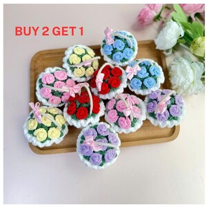 May include: A collection of crocheted flower baskets in various colors, including red, pink, yellow, blue, and purple. Each basket is adorned with a matching ribbon bow. The text "BUY 2 GET 1" is visible in the upper left corner.