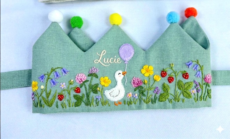 May include: A light green fabric crown with embroidered flowers, strawberries, and a white duck holding a purple balloon. The name "Lucie" is embroidered above the duck. The crown has six pom-poms in white, green, yellow, blue, and red.