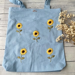 Puede incluir: Una bolsa de lona azul claro con girasoles bordados. La bolsa tiene dos asas y la palabra "Li" está bordada en la esquina superior izquierda.
