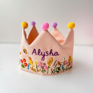 May include: A light pink fabric crown with embroidered floral details and the name "Alysha" in purple script. The crown has yellow, pink, and purple pom-poms at the points.
