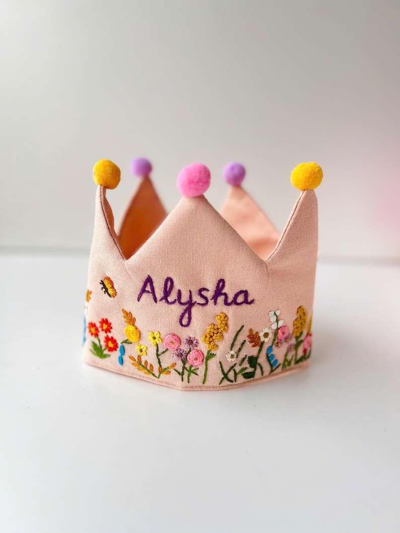 May include: A light pink fabric crown with embroidered floral details and the name "Alysha" in purple script. The crown has yellow, pink, and purple pom-poms at the points.
