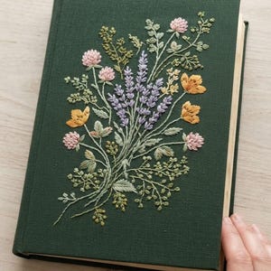 May include: A dark green hardcover book featuring an intricate floral embroidery design on the front cover. The embroidery depicts a bouquet of lavender, pink clover, and yellow wildflowers with green stems and leaves stitched onto the fabric.