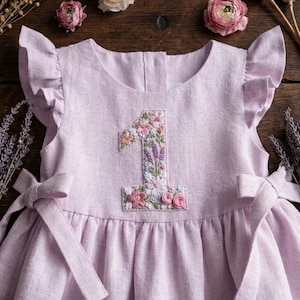 May include: A light purple linen dress with a floral embroidered number 1 on the front. The dress features ruffled sleeves, a gathered skirt, and bow ties at the waist. The dress is designed for a baby's first birthday.