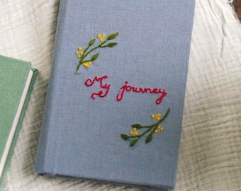 May include: A blue linen journal with the words "my journey" embroidered in red thread. The cover is decorated with two small sprigs of yellow flowers embroidered in green thread.