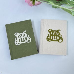 Op de afbeelding: Twee kleine hardcover boeken. Eén is olijfgroen met een wit geborduurd ontwerp, de andere is gebroken wit met een groen geborduurd ontwerp. De ontwerpen lijken gestileerde initialen of een monogram te zijn.