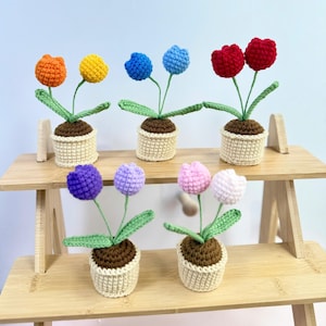 May include: A collection of six crochet tulip plants in various colors, including orange, yellow, blue, red, purple, and pink. Each tulip has green leaves and is potted in a beige crochet pot with brown soil. The plants are displayed on a wooden shelf.