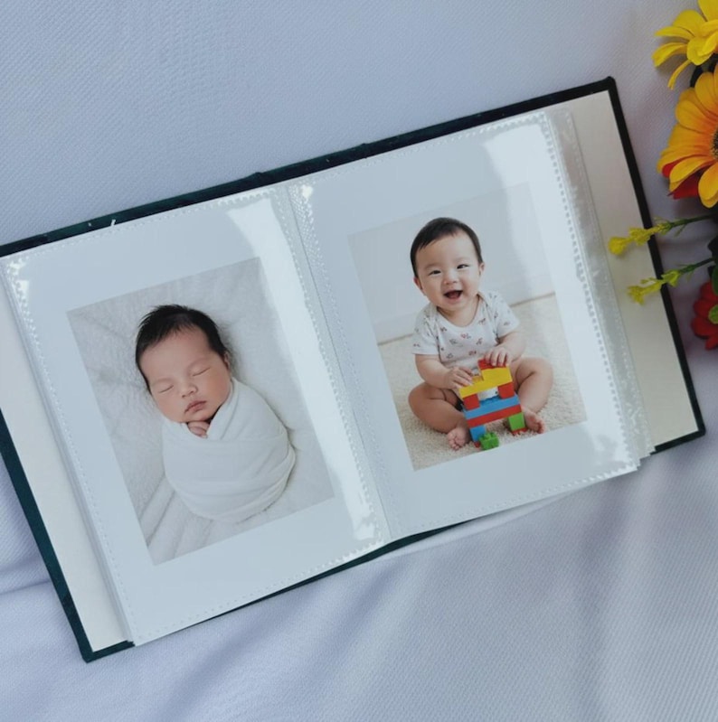 Puede incluir: &Aacute;lbum de fotos abierto con dos fotos de beb&eacute;. Una muestra a un reci&eacute;n nacido durmiendo envuelto en una manta blanca. La otra muestra a un beb&eacute; sonriente jugando con bloques de colores. El &aacute;lbum tiene una cubierta verde oscuro.