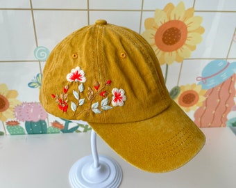 May include: A mustard yellow baseball cap with embroidered white and red flowers.