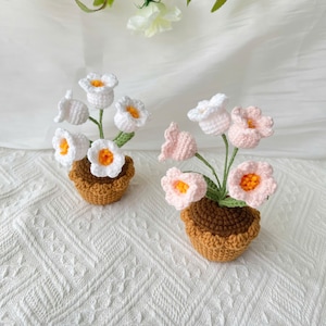 May include: Two crochet flower arrangements in small brown pots. One arrangement features white flowers with orange centers, the other has pink flowers with orange centers. The flowers are on green stems.