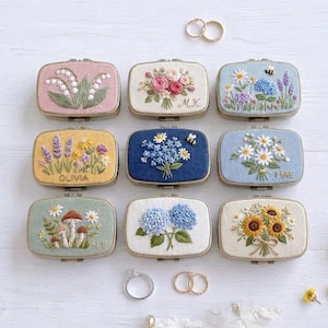 May include: Nine rectangular jewellery boxes featuring embroidered floral designs including lilies, roses, lavender, daisies, sunflowers, and hydrangeas. Some boxes include embroidered names like Olivia, M.K., and Mae, alongside small bee motifs.