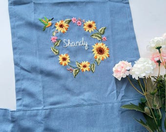 May include: A blue denim apron with a ruffled hem and adjustable straps. The apron features an embroidered sunflower wreath with the name "Shandy" in the centre.