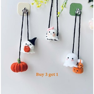 May include: Three crocheted ghost ornaments with different facial expressions and accessories, including a pumpkin, a witch's hat, and a pink bow. The ghosts are hanging from black cords attached to hooks on a white wall. The text "Buy 3 get 1" is written below the ornaments.