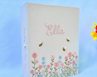 May include: A rectangular photo album with a light beige fabric cover featuring embroidered pink text that reads Ella. The cover is decorated with embroidered pink and blue wildflowers and three small yellow and black bumblebees.