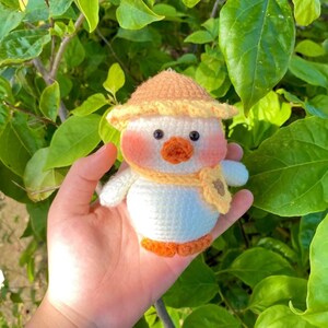 May include: A crocheted stuffed toy duck wearing a yellow and orange hat and a yellow scarf. The duck has a white body, orange feet, and a pink face.