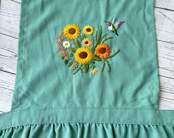 May include: A teal blue apron with a ruffled hem and embroidered sunflowers, daisies, and a hummingbird.