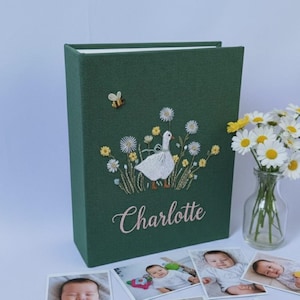 Puede incluir: Un &aacute;lbum de fotos verde oscuro con un dise&ntilde;o bordado de un ganso blanco en un campo de flores, una abeja y el nombre "Charlotte". Varias fotos de beb&eacute; est&aacute;n frente al &aacute;lbum, y un jarr&oacute;n de margaritas a la derecha.