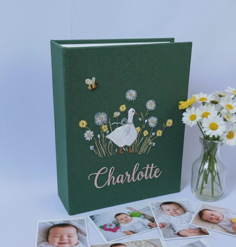 Puede incluir: Un &aacute;lbum de fotos verde oscuro con un dise&ntilde;o bordado de un ganso blanco en un campo de flores, una abeja y el nombre "Charlotte". Varias fotos de beb&eacute; est&aacute;n frente al &aacute;lbum, y un jarr&oacute;n de margaritas a la derecha.
